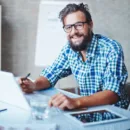 Man writing background Motivational Quote | Watertown Dentistry Smiling man with glasses and a beard seated at a bright desk, writing with a pen beside a tablet.